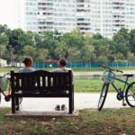 man and woman sitting on bench near two bikes viewing green field during daytime